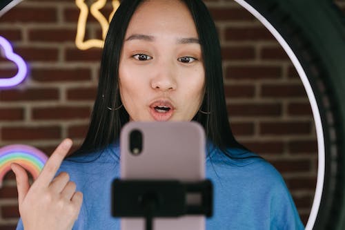 A person gestures while facing a mobile phone in a standard, and being lit by a ring light. Image by Ivan Samkov on Pexels.